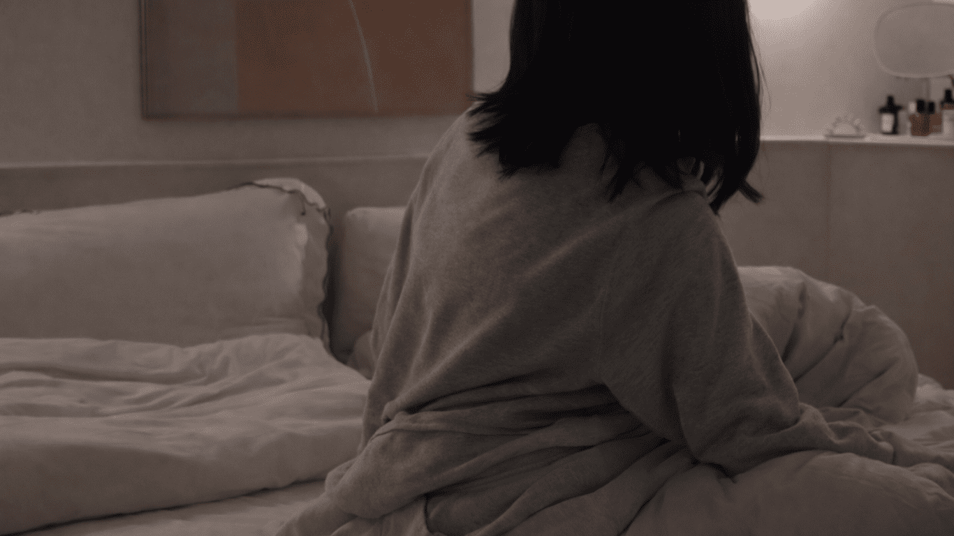 高級枕と整体枕の違いを考えながら、起床後にベッドで座る人物の後ろ姿